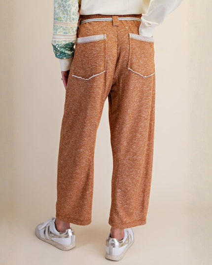 A person shown from the back wearing wide-leg cropped trousers in a heathered rust-orange color, paired with white sneakers and a cream top. The photo is shot against a solid, warm beige studio background.