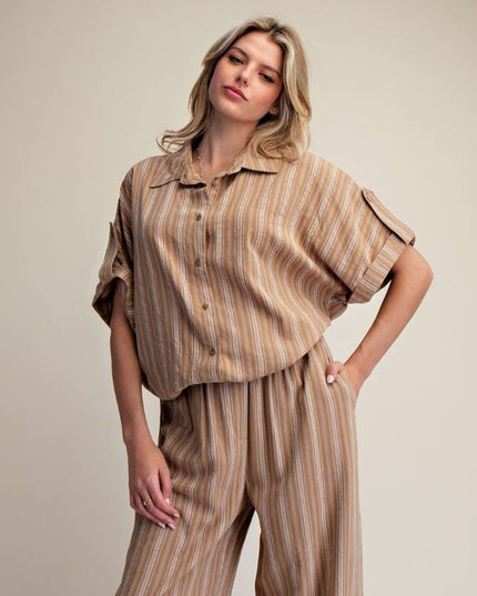 A woman modeling a matching two-piece set consisting of a short-sleeve button-up shirt and wide-leg trousers, both in a beige and brown vertical stripe pattern, against a plain beige studio background.