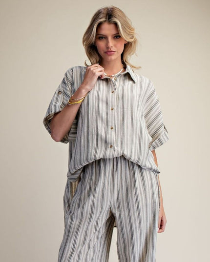 A woman modeling a matching short-sleeve button-up shirt and wide-leg trousers set in a light gray and white vertical stripe fabric, photographed against a solid beige studio background.