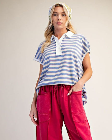 A woman modeling a short-sleeve polo shirt with blue and white horizontal stripes, paired with bright pink cargo pants and a patterned headscarf, against a plain beige studio background.
