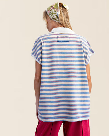 A woman shown from the back wearing a short-sleeve polo shirt with horizontal blue and off-white stripes, paired with magenta pants and a floral headscarf, against a solid light beige background.
