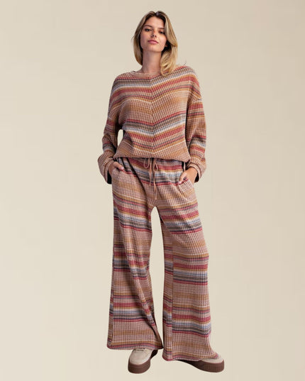 A woman modeling a matching two-piece lounge set featuring a long-sleeve top and wide-leg pants in a textured waffle knit with a multicolor horizontal stripe pattern of beige, brown, red, and blue, set against a solid beige background.