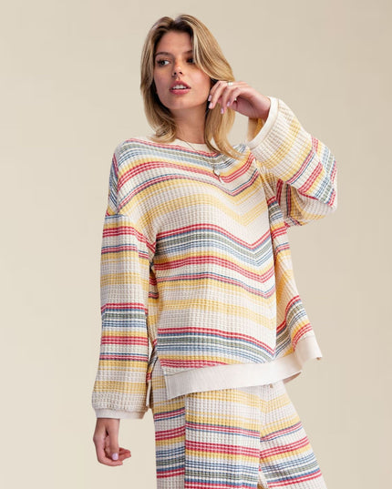 A woman modeling a matching two-piece waffle-knit loungewear set featuring a long-sleeve top and pants in a multicolor chevron stripe pattern of yellow, red, blue, and gray on a cream base, set against a solid beige studio background.