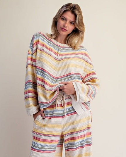 A woman modeling a matching two-piece lounge set with a waffle-knit texture and horizontal multicolor stripes in yellow, blue, red, and gray on a cream base, against a solid beige background.