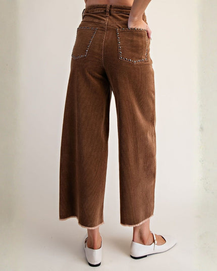 A person shown from the back wearing brown corduroy wide-leg cropped pants with studded pocket details, paired with white Mary Jane flats, against a plain off-white studio background.