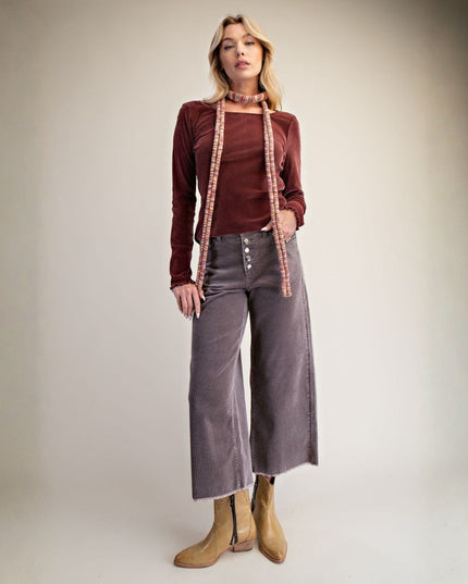 A woman modeling a long-sleeve burgundy velvet top with a decorative pink and gold patterned collar trim, paired with wide-leg mauve corduroy pants and tan ankle boots, against a neutral studio backdrop.