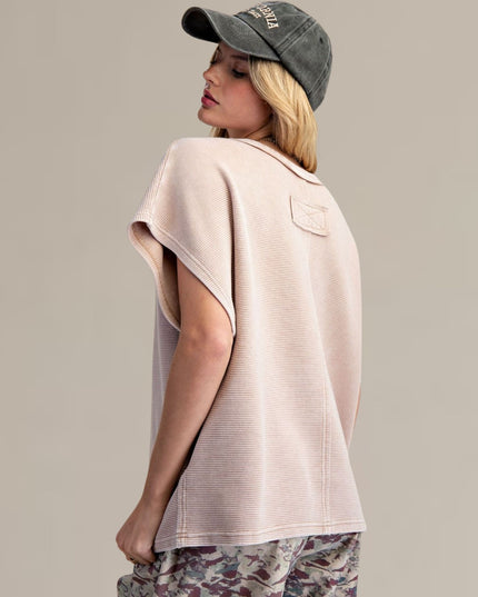 A woman shown from the back wearing a light blush pink ribbed knit t-shirt with a small back pocket, a dark gray baseball cap, and camouflage pants, set against a solid light gray background.