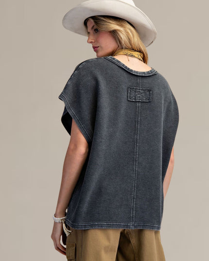A woman shown from the back wearing a dark washed denim short-sleeve top with a patch pocket, tan pants, and a white cowboy hat against a solid beige studio background.