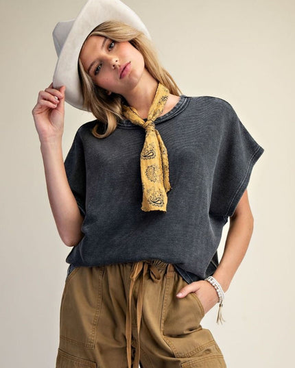 A woman wearing a dark charcoal gray short-sleeve top, a yellow paisley neck scarf, and brown cargo pants, holding a white cowboy hat against a plain beige studio background.