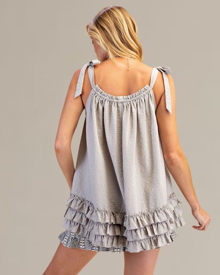 A woman shown from the back wearing a light gray sleeveless tie-shoulder top with a ruffled tiered hem, photographed against a plain warm gray studio background.