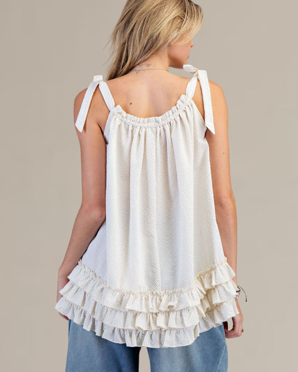 A woman shown from the back wearing a white sleeveless ruffled top with tie straps and light blue jeans, against a plain beige studio background.