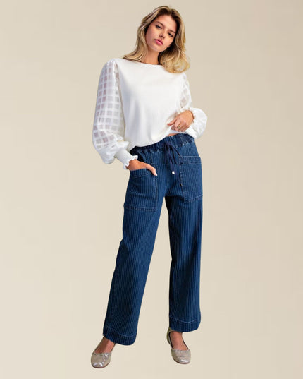 A woman modeling a white long-sleeve top with sheer patterned puff sleeves and dark blue wide-leg pinstripe jeans, paired with metallic flats, against a solid beige background.