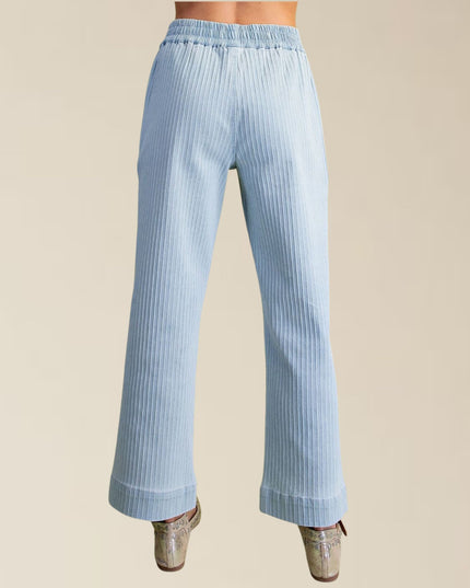 Light blue pleated wide-leg pants shown from the back on a model, paired with metallic silver heels, against a solid beige background.