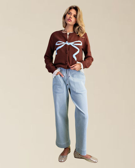 A woman modeling a brown button-up cardigan with a light blue ribbon graphic on the front, paired with light blue ribbed wide-leg jeans and metallic flats, set against a solid beige background.