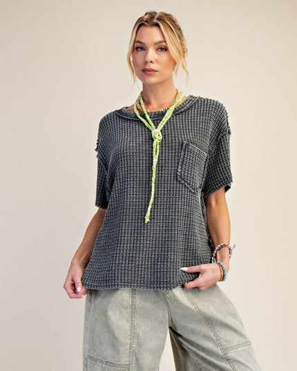 A woman modeling a short-sleeve, oversized black and white houndstooth top with a chest pocket and a neon green knotted scarf, paired with light gray cargo pants against a solid beige background.