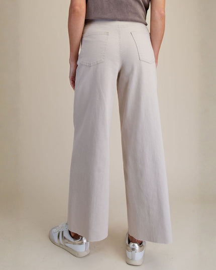 A person shown from the back wearing light beige wide-leg trousers, a taupe top, and white sneakers against a warm beige studio background.