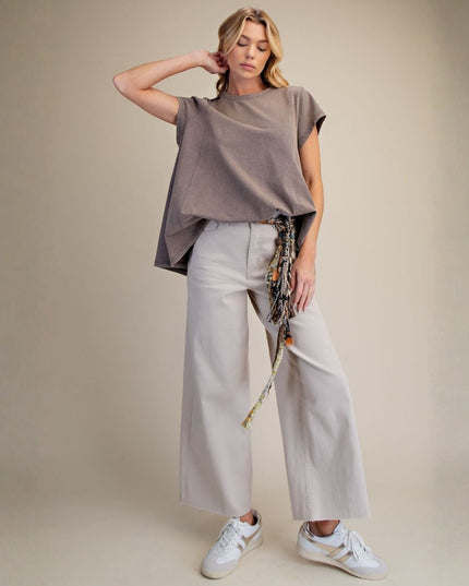 A woman models a casual outfit consisting of a taupe short-sleeve top, light beige wide-leg trousers, and white sneakers, styled with a patterned belt against a neutral beige studio background.