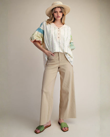 A woman stands against a plain beige studio background wearing a cream patterned short-sleeve blouse with colorful puff sleeves, beige wide-leg pants, a straw hat, and green slide sandals.