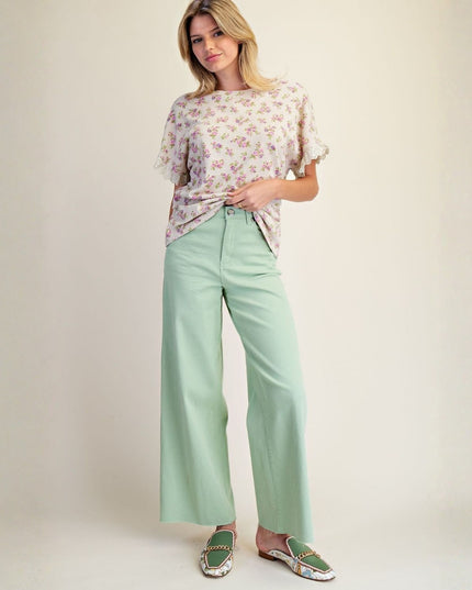 A woman modeling a light green wide-leg pants outfit with a short-sleeve floral top and green patterned slip-on shoes, set against a plain beige studio background.