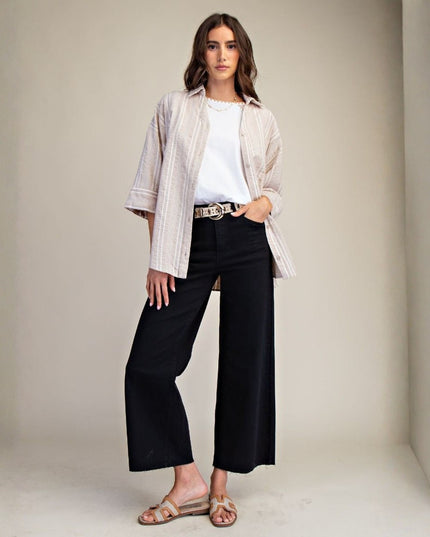 A woman stands in a studio wearing a light beige striped button-up shirt over a white T-shirt, black wide-leg cropped pants, and tan slide sandals.
