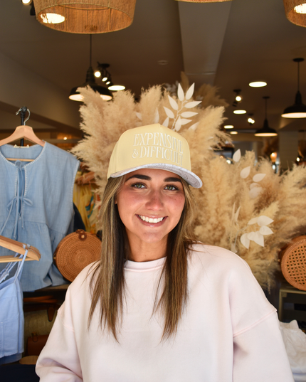 Woman wearing a beige cap and white sweatshirt in a store setting with clothing and decor in the background.