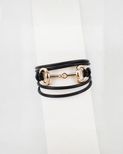 A multi-layered wrap bracelet featuring black leather cords and a polished gold-tone horsebit clasp, displayed on a white cylindrical stand.