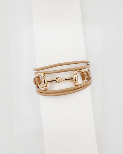 A multi-layered wrap bracelet featuring tan leather bands and polished gold-tone horsebit hardware, displayed on a white cylindrical stand.