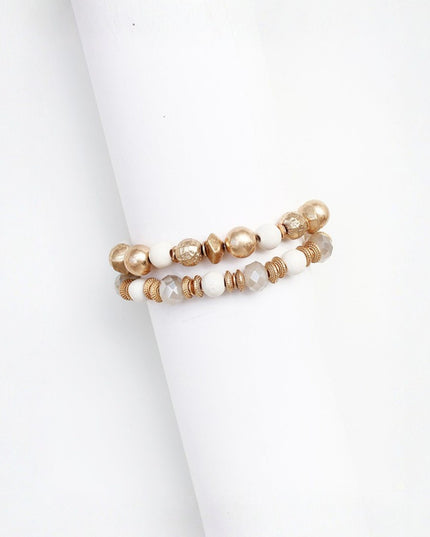 Two beaded stretch bracelets displayed on a white cylindrical stand. The bracelets feature a mix of white beads, metallic gold beads, and faceted clear beads with a warm gold-toned setting.