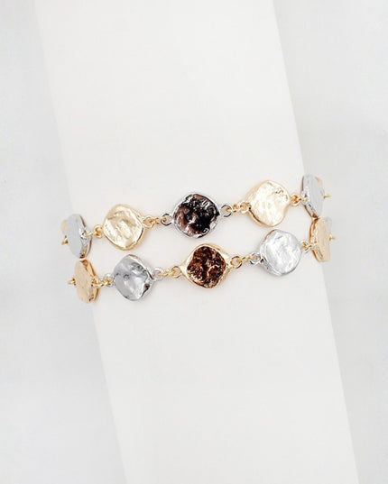 Two delicate bracelets displayed on a white cylindrical stand. One bracelet is gold-toned with textured round and square metallic charms, and the other is rose gold-toned with clear faceted stones.