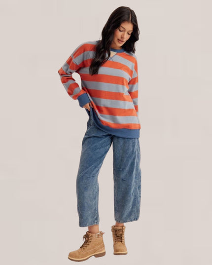 A woman modeling a striped crewneck sweatshirt with orange and gray horizontal stripes, paired with wide-leg blue jeans and tan lace-up boots, against a solid light beige background.