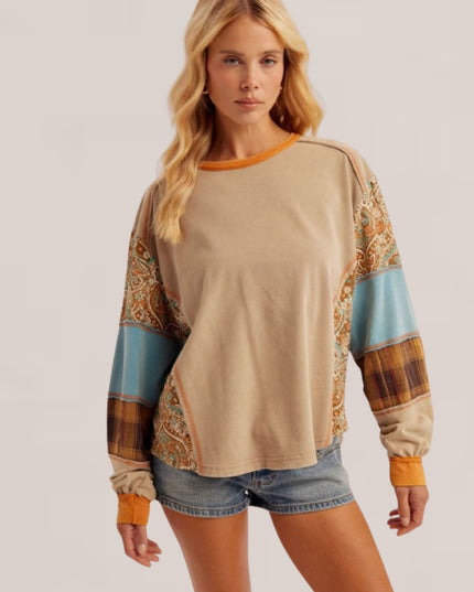 A woman modeling a beige long-sleeve top with orange trim and multicolor patterned color-blocked sleeves, paired with denim shorts, against a light gray studio background.