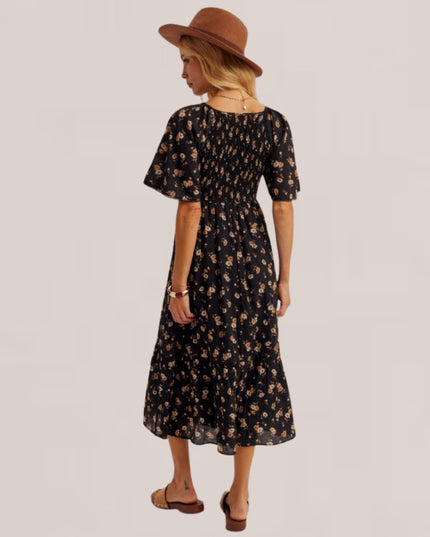 A woman shown from the back wearing a black midi dress with a small beige floral print, a brown wide-brim hat, and brown slide sandals against a light gray studio background.