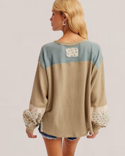 A woman shown from the back wearing a color-block waffle-knit sweater with a dusty blue upper section, a beige lower section, and floral lace trim on the sleeves and back pocket, set against a plain light gray studio background.