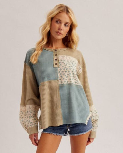 A woman modeling a patchwork knit sweater with beige, light blue, and floral patterned sections, paired with denim shorts, against a plain light gray studio background.