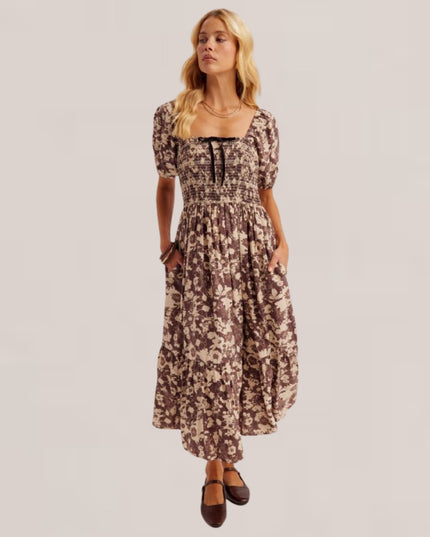 A woman modeling a brown and beige floral print midi dress with puff sleeves, a smocked bodice, and a square neckline, set against a solid light gray background.
