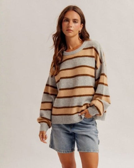 A woman modeling a cozy knit sweater with horizontal stripes in beige, brown, and light gray, paired with denim shorts against a plain light gray background.