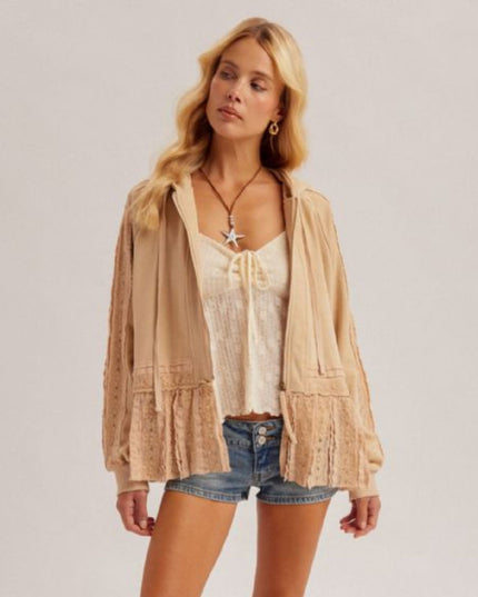 A woman modeling a beige lace-trimmed open-front cardigan over a cream lace-trimmed camisole and denim shorts, photographed against a plain light gray studio background.