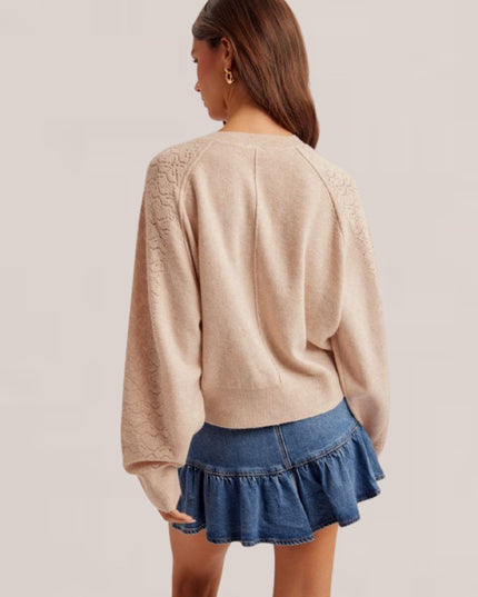 A woman shown from the back wearing a light beige knit sweater with lace-like detailing on the shoulders and a blue denim ruffled mini skirt, against a plain light beige studio background.