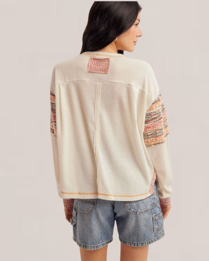 A woman shown from the back wearing a cream-colored knit sweater with multicolor patterned knit sleeve panels and a matching patch on the upper back, paired with denim shorts against a light gray studio background.