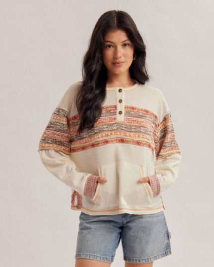 A woman modeling a cream-colored knit sweater with horizontal multicolor striped patterns across the chest and sleeves, paired with denim shorts, against a light gray studio background.
