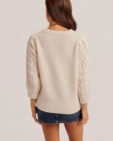 A woman shown from the back wearing a cream-colored cable-knit sweater with textured balloon sleeves and a dark denim mini skirt, set against a plain light beige studio background.