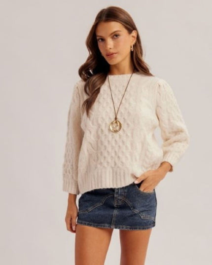 A woman modeling a cream cable-knit sweater with puffed sleeves, paired with a blue denim mini skirt and a gold pendant necklace, against a light gray studio background.