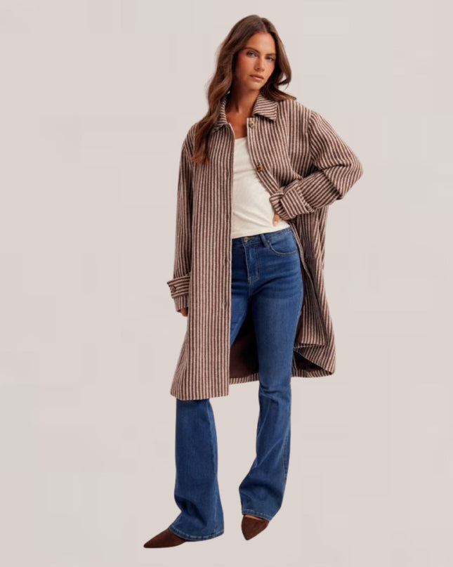 A woman modeling a long brown and beige vertically striped button-up shirt jacket over a white top and blue flared jeans, photographed against a plain light beige studio background.