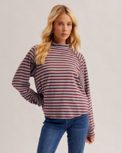 A woman modeling a long-sleeve knit top with horizontal red and gray stripes, featuring textured knit shoulder panels and a mock neck, paired with blue jeans against a light gray studio background.