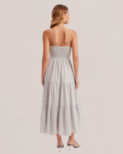 A woman viewed from the back wearing a sleeveless, tiered maxi dress with thin straps and a smocked bodice, paired with black strappy heels, against a light beige studio background.