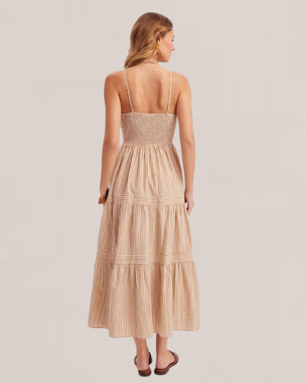 A woman viewed from the back wearing a sleeveless, smocked, tiered maxi dress in a light beige or cream color with thin vertical stripes. She is standing against a solid, light warm gray/beige studio background.