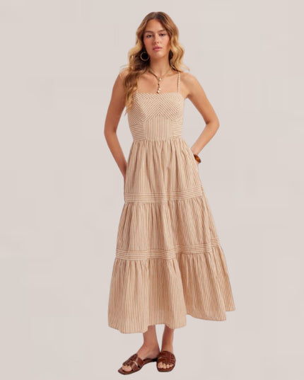 A woman modeling a beige and cream striped tiered maxi dress with crochet detailing on the bodice, paired with brown leather slide sandals, against a solid light beige background.