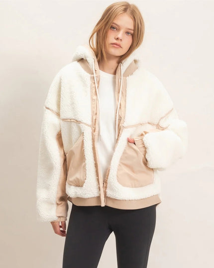 A woman wearing a cream and beige sherpa fleece zip-up hoodie jacket with a hood, layered over a white top and black pants, standing against a plain off-white studio background.