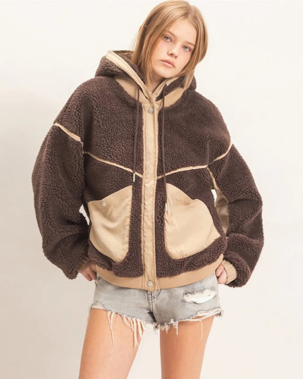 A model wearing a brown and beige sherpa fleece zip-up hoodie jacket with large front pockets, paired with distressed denim shorts, against a plain off-white background.