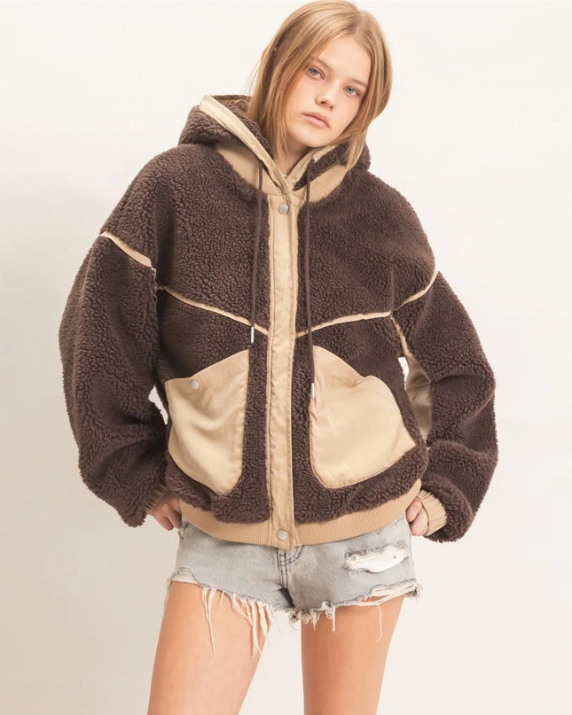 A model wearing a brown and beige sherpa fleece zip-up hoodie jacket with large front pockets, paired with distressed denim shorts, against a plain off-white background.
